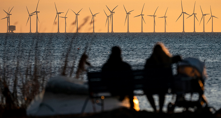 Två personer sitter på en bänk framför havet. Ute i havet syns vindkraftverk.