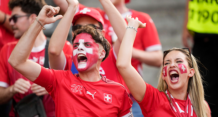 En man och en kvinna står på läktaren och skriker. Det är en match i fotboll. De har målat Schweiz flagga i ansiktet.