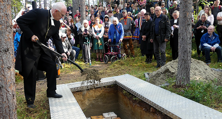 Han står vid en grav. Han häller ner jord med en spade. Bakom syns många människor.