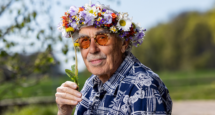 Han har en blomsterkrans på huvudet. Han har glasögon och mustasch. Han håller i en maskros.