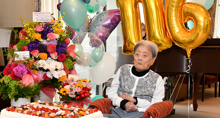 Hon sitter bland massa blommor. Bakom henne är guldiga ballonger som visar siffran 116.