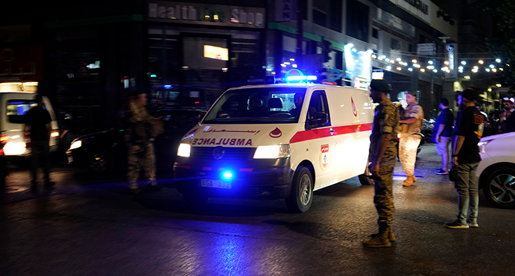 En ambulans med blåljus kör på en gata. Det står militärer vid sidan.