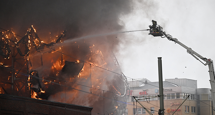 Det brinner i en stor byggnad. En kran är högt uppe i luften. I kranens korg står en brandman och sprutar vatten på elden.