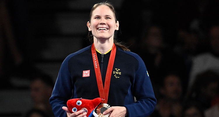 Hon står med medalj om halsen. Hon håller i medaljen och i ett rött gosedjur. Hon är glad.