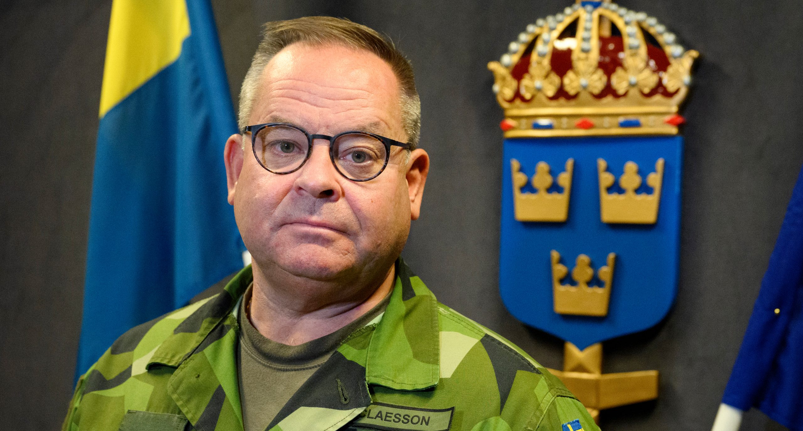 Han har gröna militärkläder. Han har glasögon, Han står framför en svensk flagga och en skylt med tre kronor.