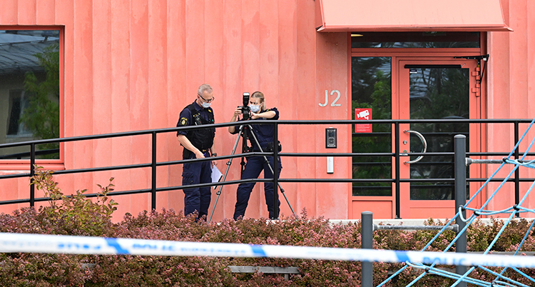 Två poliser står framför skolans röda vägg. De ställer in en kamera.