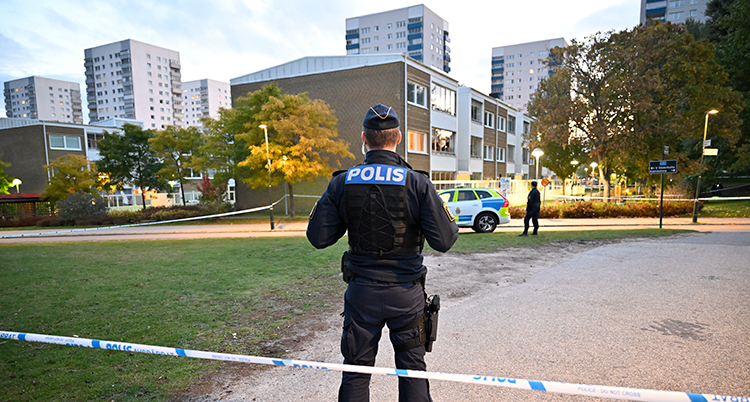 En polis står bakom en avspärrning. Vi ser hans rygg. På den står det polis. Framör honom är flera hus.