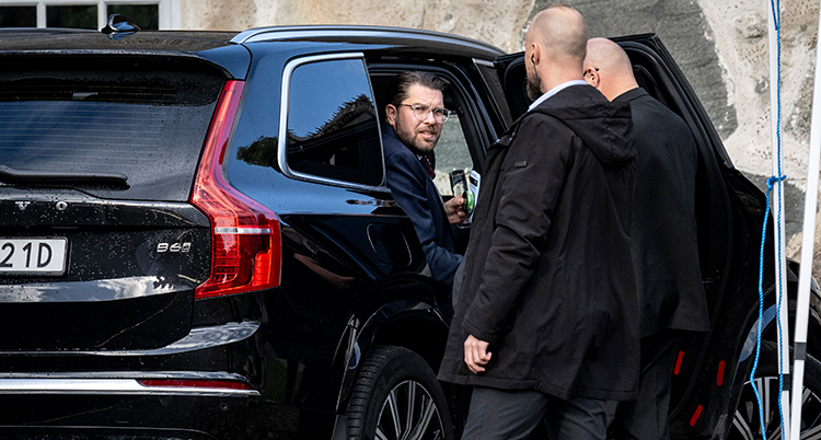 Han sitter i en svart bil med öppen dörr. Två personer i mörka rockar står vid honom.