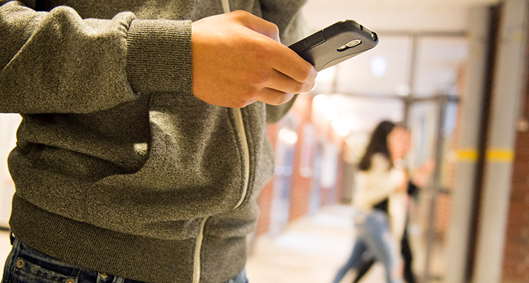 En ung person står i en skolkorridor och tittar på en mobil. Vi ser bara personens torso och hand som håller i mobilen.