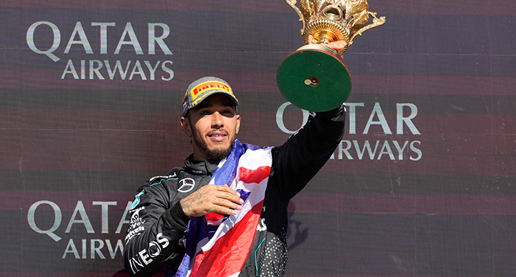 Han håller upp en guldpokal. Runt axlarna har han en brittisk flagga. Han har sitt lags kläder på sig, Mercedes.