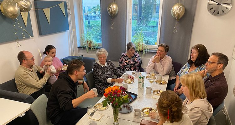 Många människor sitter runt ett bord. Det finns tårta och blommor på bordet.