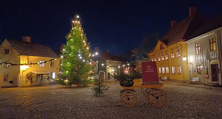 En stor gran står på torget. Runt torget finns hus i trä. Det lyser i fönstren.