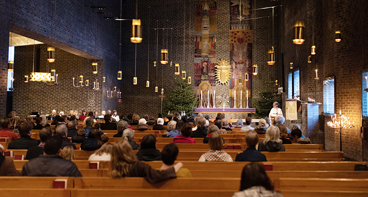 Människor sitter i bänkar i kyrkan. En präst står längst fram och pratar.