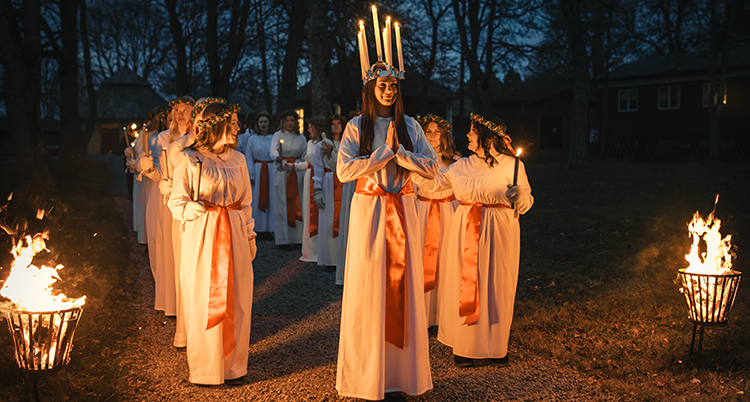 Lucia har en krona med ljus på huvudet. Bredvid henne går tärnor. Alla har vita klänningar och ett rött band i midjan.