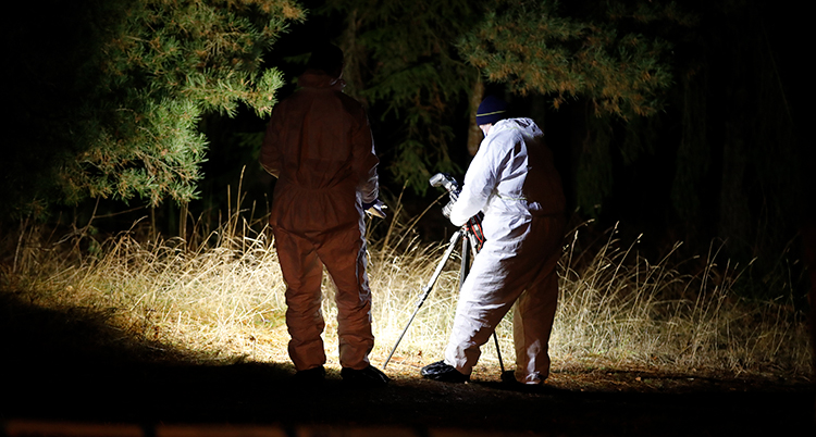 Två personer i ljusa overaller jobbar i mörkret. En lyser med en lampa, den andre har en kamera på ett stativ.