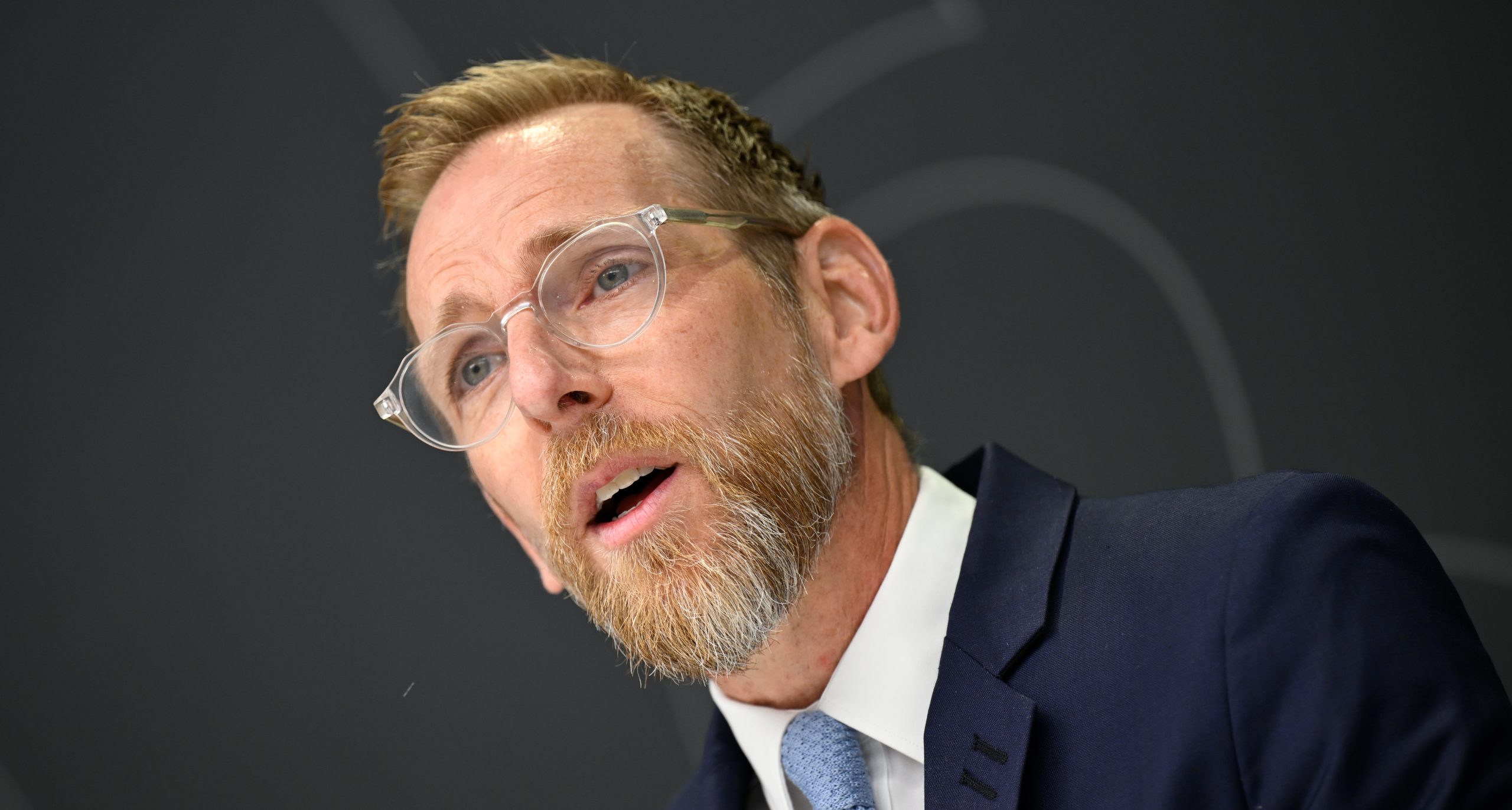 Han har glasögon och skägg. Han pratar på en presskonferens.