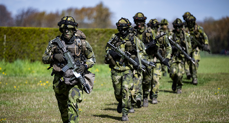 Militärer i gröna kläder går på rad. De har hjälmar och vapen.