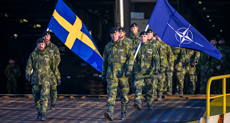 Led med soldater går på en ramp på ett fartyg. De bär på två flaggor, en svensk och en Natoflagga.