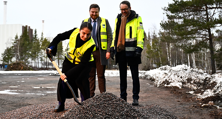 Hon gräver med en spade. Två män står bakom. Längre bort syns et kärnkraftverk.