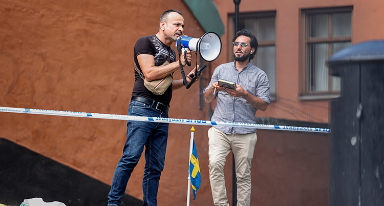 Den ena mannen har svarat t-shirt. Han håller i en megafon. Den andra har en vit skjorta. Han håller i en koran.