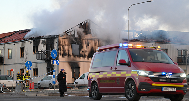 En bil från räddningstjänsten står längst fram i bilden. Bakom syns huset som brinner. Det är mycket rök. Delar av huset är förstört.