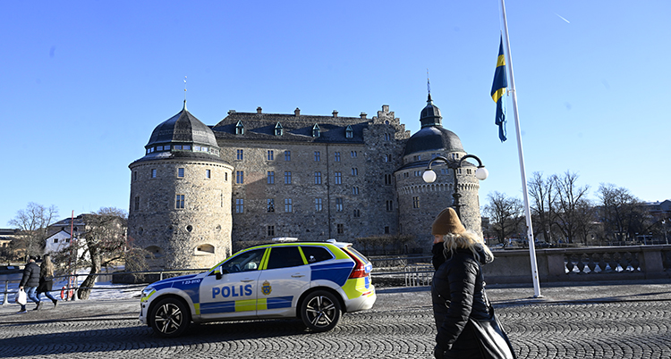 Slottet i bakgrunden. En flaggstång med en flagga på halva stången. En polisbil och en människa som går.