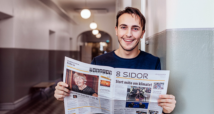 Han håller i en tidning. Han står lutad mot en vägg.