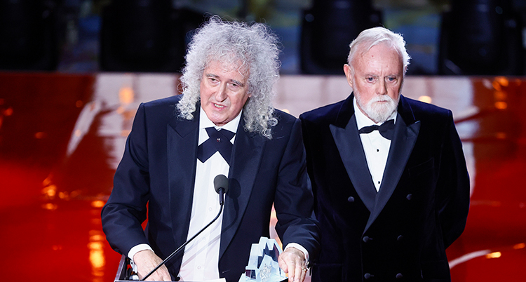 De står på en scen. De har festkläder på sig. Brian May pratar i en mikrofon.