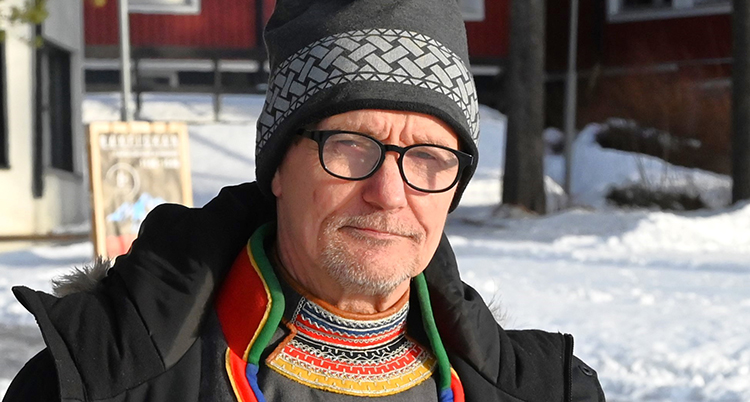Han har glasögon och mössa. Han tittar in i kameran. Han står ute. Det är snö i bakgrunden.