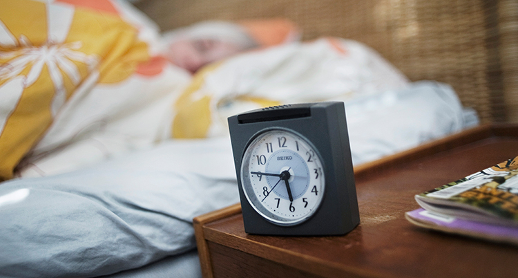 En klocka står på ett bord bredvid en säng. En kvinna sover.