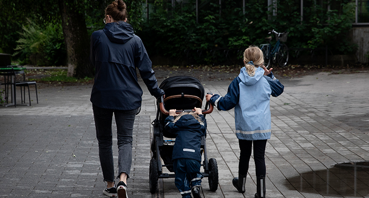 En mamma med en barnvagn och ett barn bredvid vagnen. Vi ser deras ryggar.