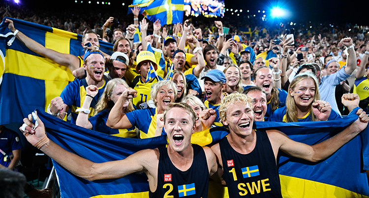 De står bredvid varandra och håller i en svensk flagga. Många personer står bakom och jublar.
