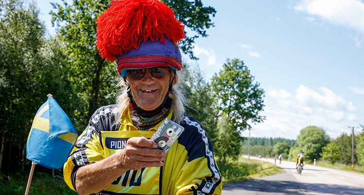 Han har en stor mössa på huvudet. Han håller i ett kassettband. Bakom syns cyklister på en väg.