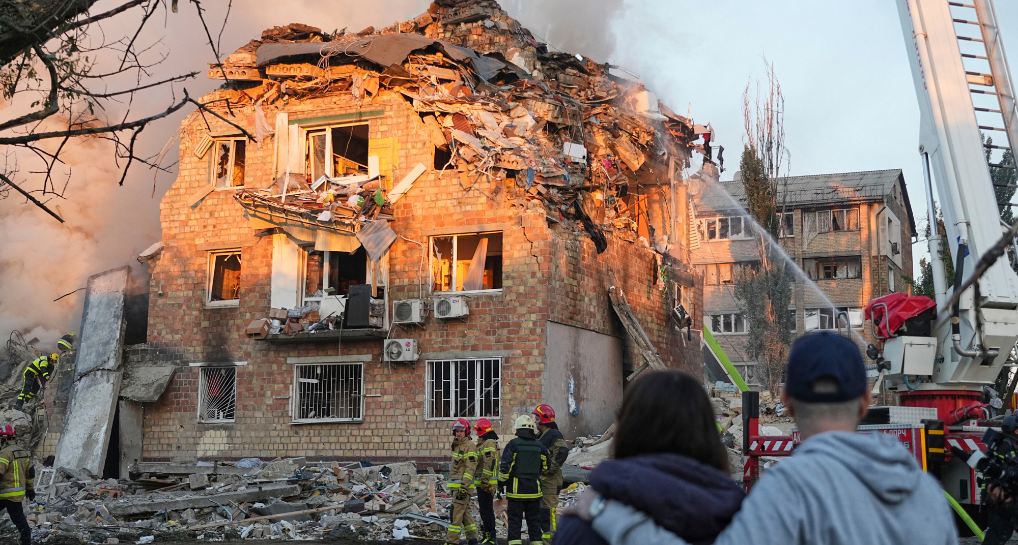 Det ryker från ett mycket skadat hus. Brandmän jobbar. Folk håller om varandra och tittar på.