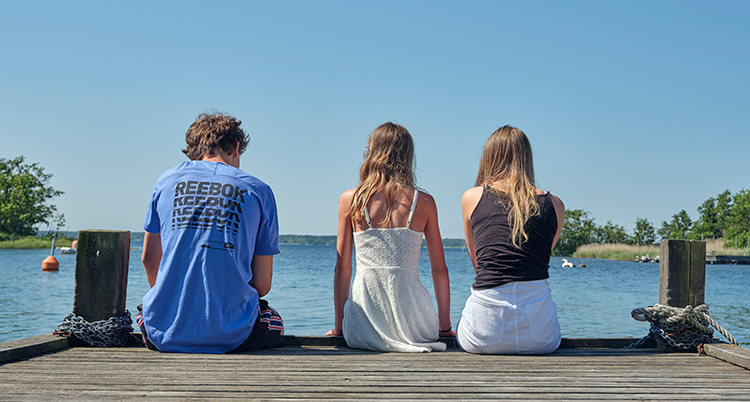 Tre unga personer sitter på en brygga och tittar ut över vattnet.