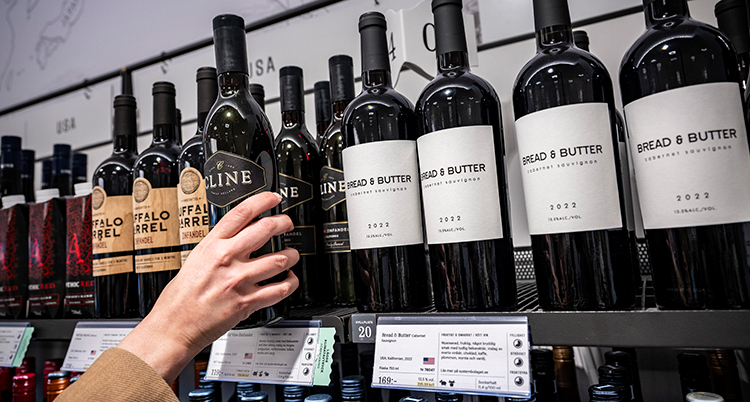 En hand tar en flaska vin på en hylla med vin på Systembolaget.