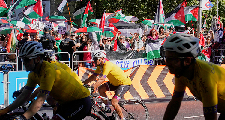 Några personer cyklar på en gata. Bredvid står många människor och protesterar. De har flaggor för Palestina.