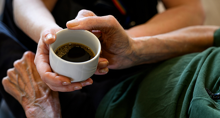En person som jobbar på ett äldreboende ger en kopp kaffe till en gammal person.