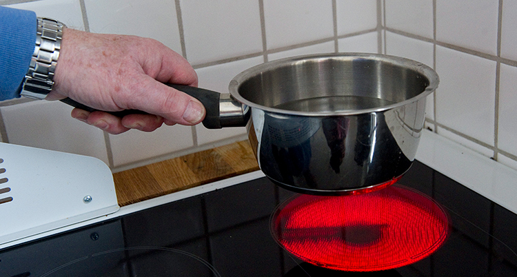En person lyfter upp en kastrull från en platta. I kastrullen finns det vatten.