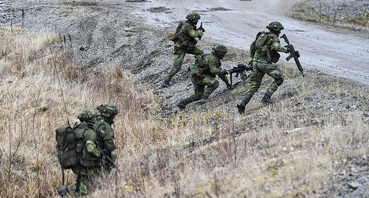 Några soldater med vapen och ryggsäckar springer på ett fält.