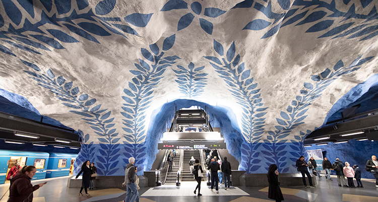 En bild från en tunnelbanestation under marken. Väggarna är målade vita och blåa. Blå löv klättrar på väggar och tak.