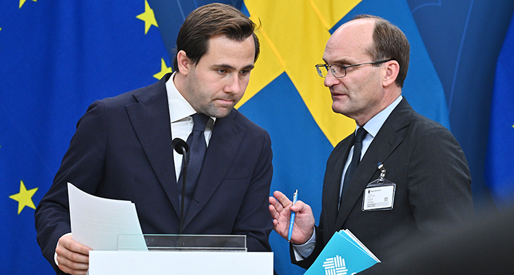 Två män står på en scen på en träff med journalister. I bakgrunden är Sveriges flagga och EUs flagga.