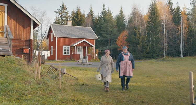 Två personer går framför ett rött hus med vita knutar.