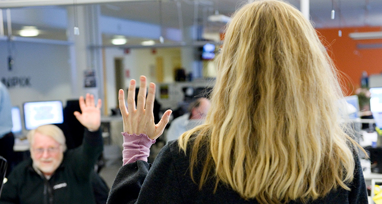 En person med långt hår står med ryggen mot kameran och hälsa med handen. En person vid ett skrivbord hejar tillbaka med handen.