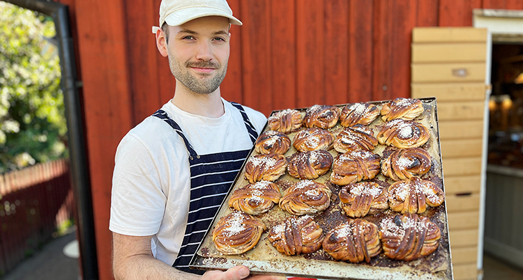 Han står framför ett rött hus, han håller fram en plåt med kanelbullar.