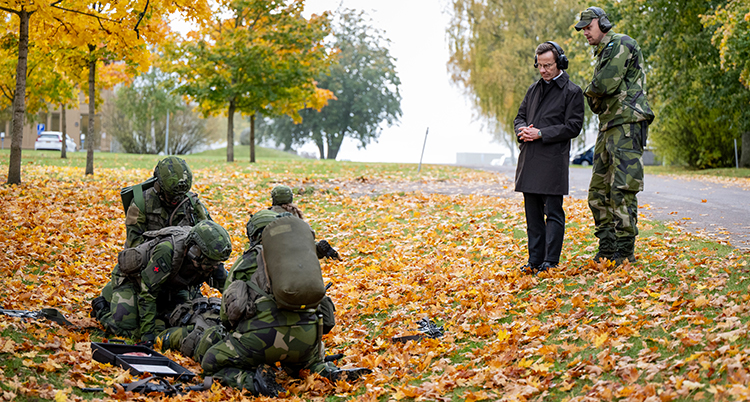 Han står framför soldater i gröna kläder. Höstlöv ligger på marken.