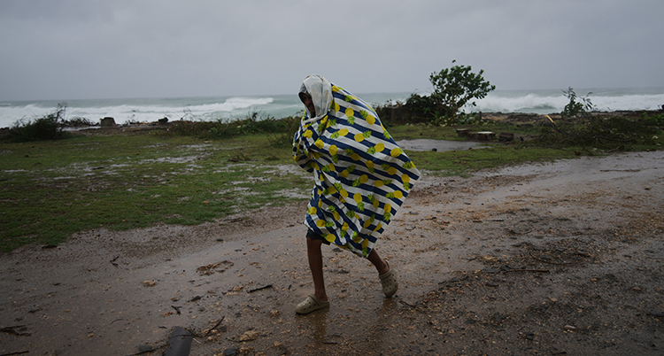 En man med ett täcke som skydd går i kraftigt regn. Bakom honom stormar ett hav.
