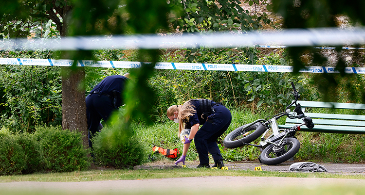 En polis är böjd över marken. Området är avspärrat med polisernas band. En cykel står lutad mot en bänk.