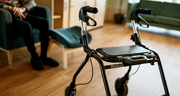 En rullator i ett rum. I bakgrunden sitter en gammal människa i en fåtölj.