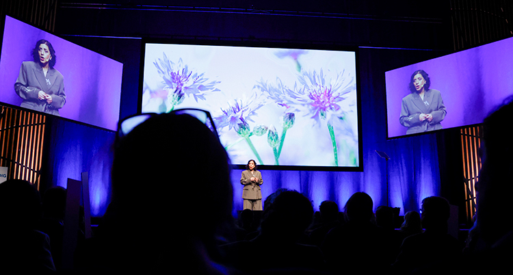 Hon står på en scen och talar. Hon syns också på två stora skärmar.
